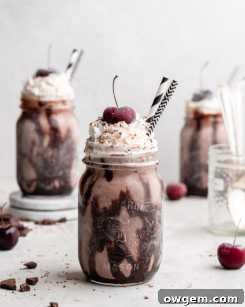 A creamy Black Forest Chocolate Cherry Milkshake in a glass, topped with a generous amount of whipped cream and a cherry, against a rustic background. Features rich chocolate ice cream and sweet cherry jam.