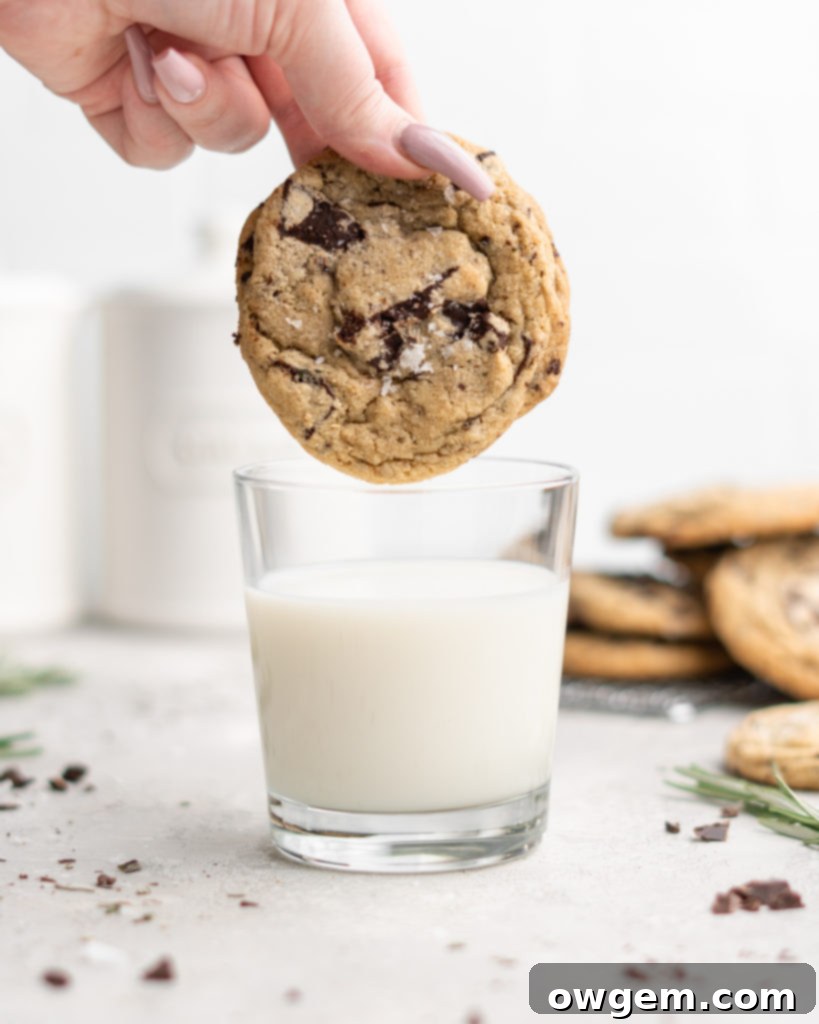 Two freshly baked Rosemary Chocolate Chunk Cookies, one showing a close-up of melted chocolate chunks and a delicate rosemary leaf on top.