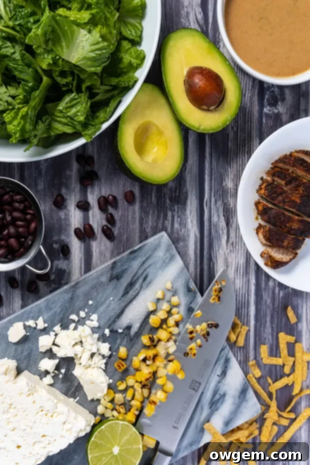 All the fresh ingredients for a Santa Fe Chicken Salad laid out: succulent Cajun chicken, vibrant romaine lettuce, golden corn kernels, rich feta cheese, wholesome black beans, sweet Medjool dates, crispy tortilla strips, ripe avocado, and bright lime wedges, awaiting their transformation into a delicious meal.