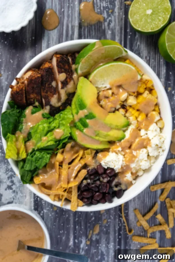 A vibrant Santa Fe Chicken Salad in a large mixing bowl, featuring Cajun chicken, fresh romaine lettuce, sweet corn, crumbled feta, hearty black beans, finely diced dates, and creamy avocado, all coated in the tantalizing peanut lime vinaigrette and topped with crunchy tortilla strips, ready to be served.